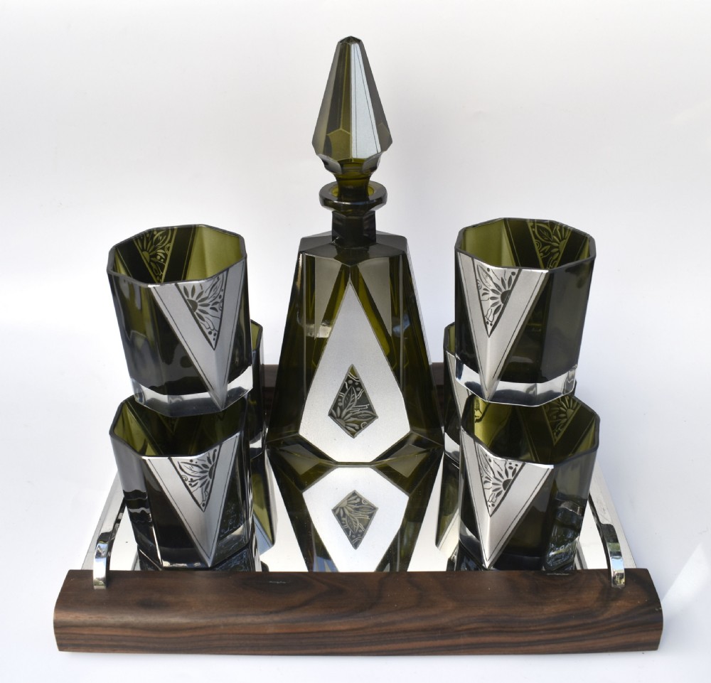 An Art Deco olive green glass decanter set featuring a faceted pyramidal bottle with a matching geometric stopper and six small angular tumblers etched with triangular patterns. View II