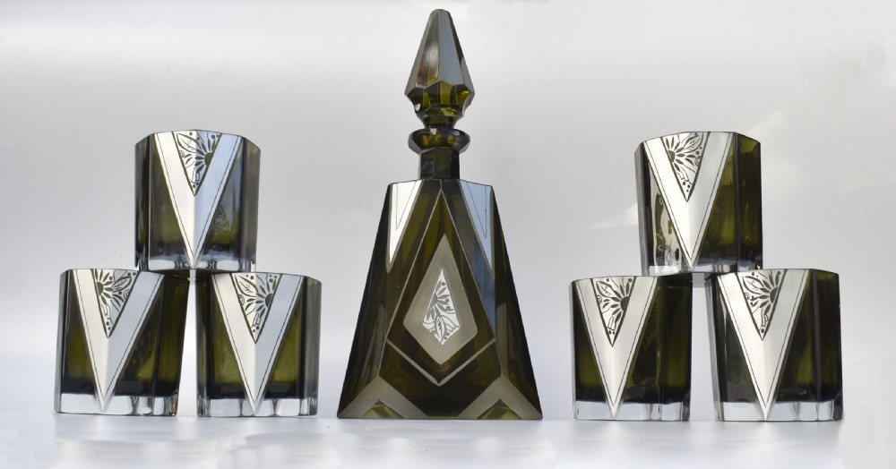 An Art Deco olive green glass decanter set featuring a faceted pyramidal bottle with a matching geometric stopper and six small angular tumblers etched with triangular patterns. View VIII