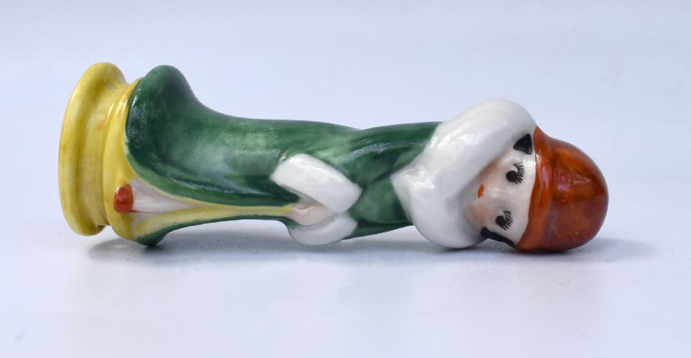 An Art Deco glazed porcelain half doll depicting a flapper in a green coat with a white fur collar, wearing an orange cloche hat and standing on a yellow base. View V