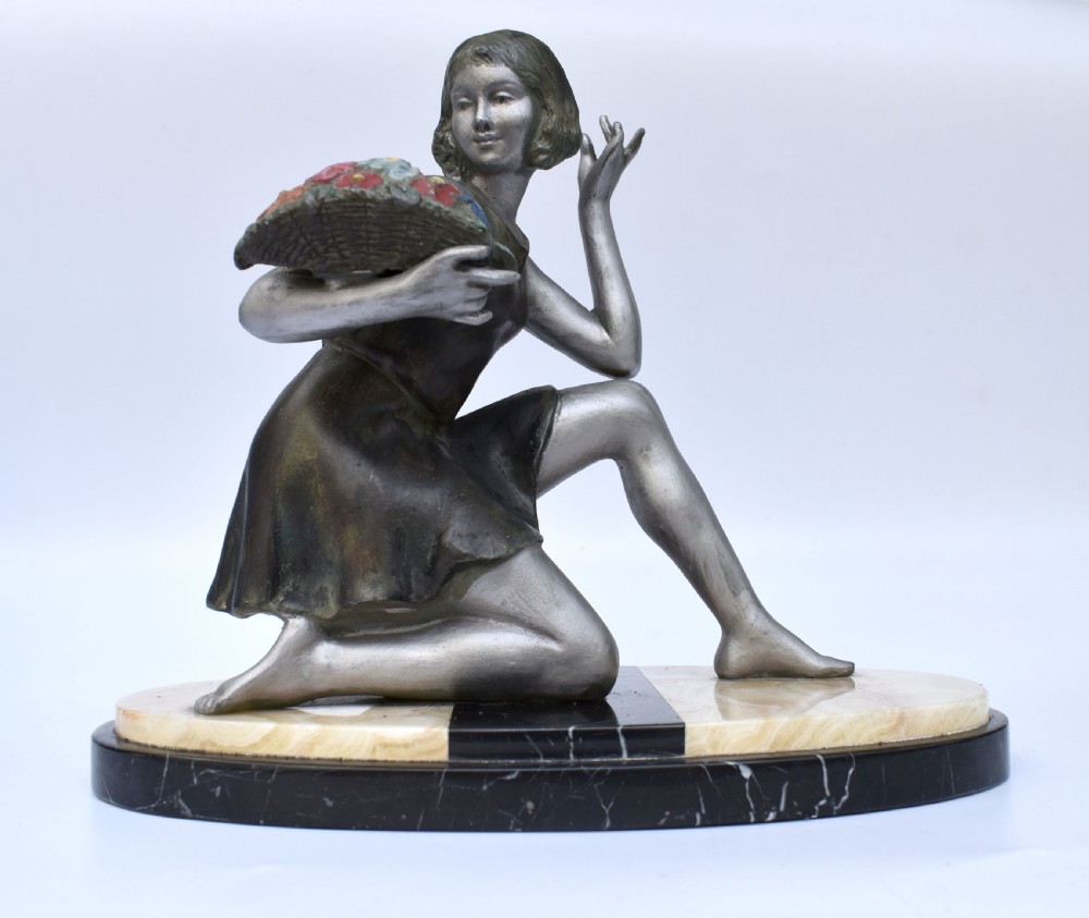 An Art Deco silver-toned spelter sculpture depicting a kneeling female figure holding a basket of coloured flowers, mounted on an oval cream onyx and black marble base.