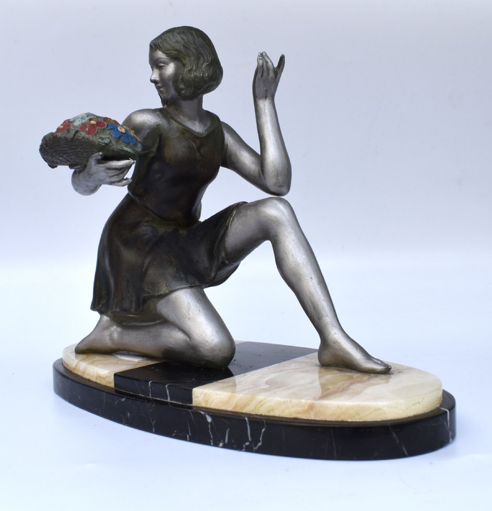 An Art Deco silver-toned spelter sculpture depicting a kneeling female figure holding a basket of coloured flowers, mounted on an oval cream onyx and black marble base. View II
