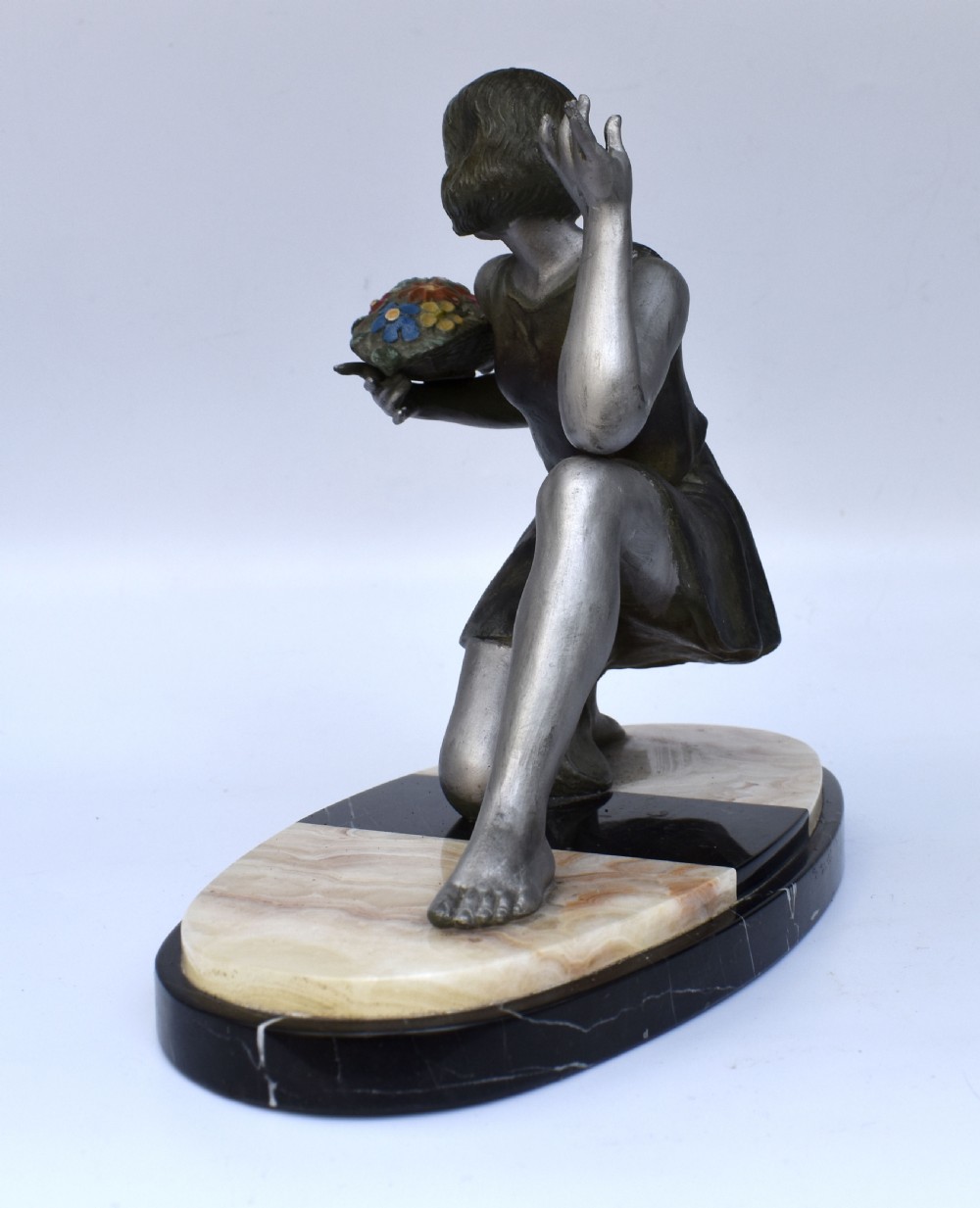 An Art Deco silver-toned spelter sculpture depicting a kneeling female figure holding a basket of coloured flowers, mounted on an oval cream onyx and black marble base. View III