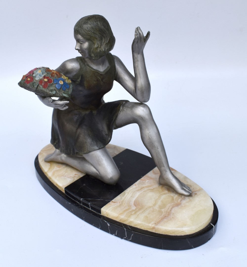 An Art Deco silver-toned spelter sculpture depicting a kneeling female figure holding a basket of coloured flowers, mounted on an oval cream onyx and black marble base. View IV