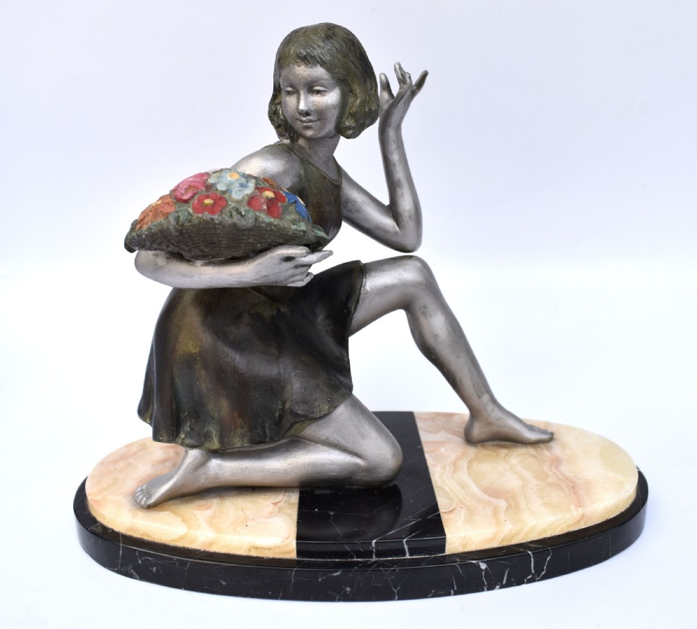 An Art Deco silver-toned spelter sculpture depicting a kneeling female figure holding a basket of coloured flowers, mounted on an oval cream onyx and black marble base. View VIII