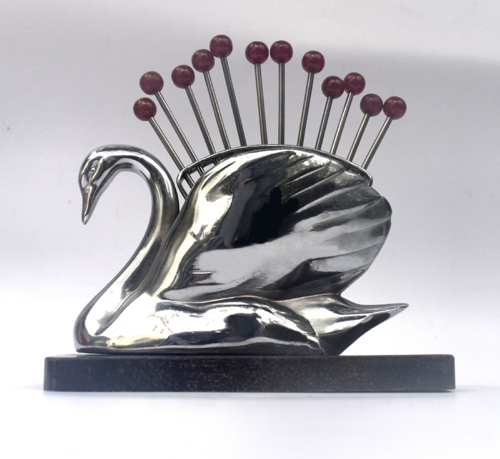 An Art Deco chrome swan holder holding red Bakelite-topped cocktail picks set upon a dark wooden base.