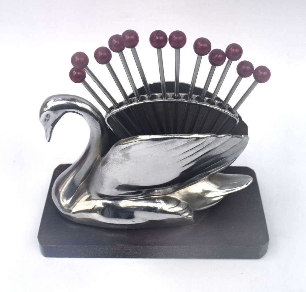 An Art Deco chrome swan holder holding red Bakelite-topped cocktail picks set upon a dark wooden base. View II