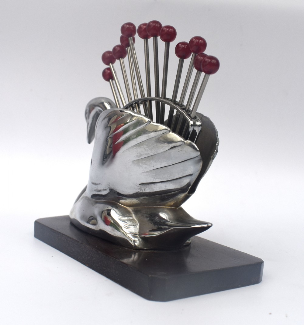 An Art Deco chrome swan holder holding red Bakelite-topped cocktail picks set upon a dark wooden base. View IV