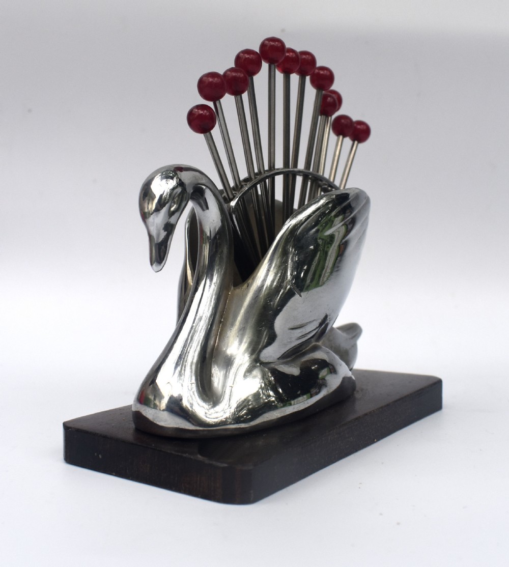 An Art Deco chrome swan holder holding red Bakelite-topped cocktail picks set upon a dark wooden base. View V