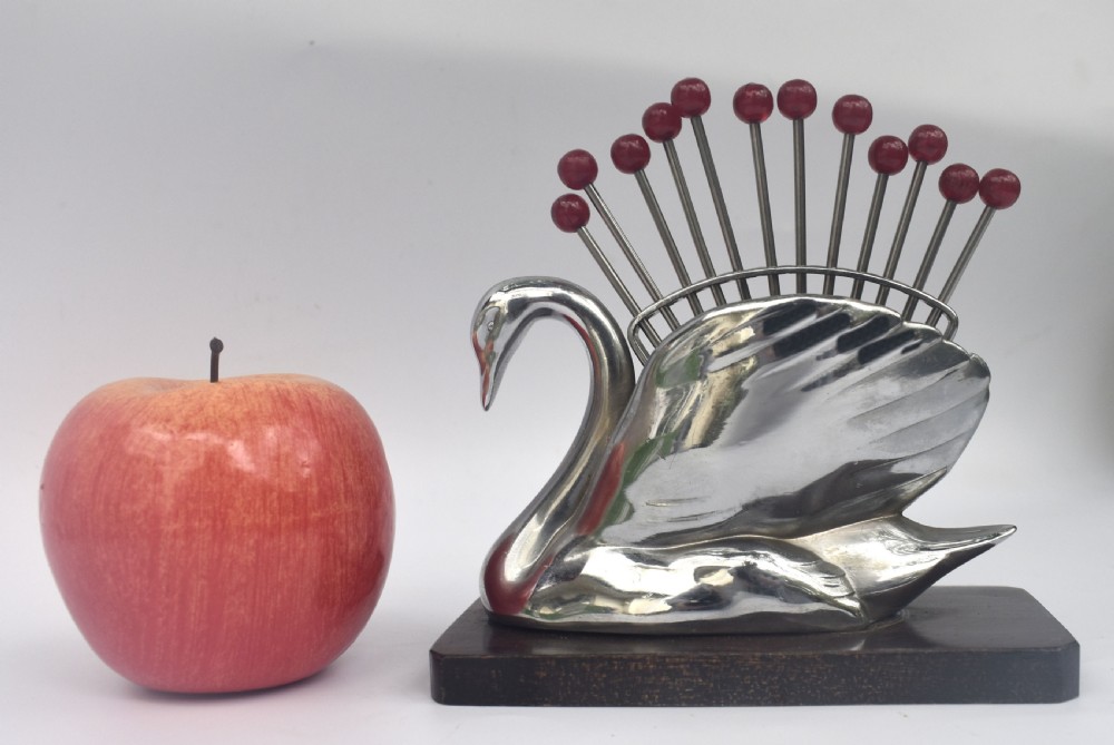 An Art Deco chrome swan holder holding red Bakelite-topped cocktail picks set upon a dark wooden base. View VI
