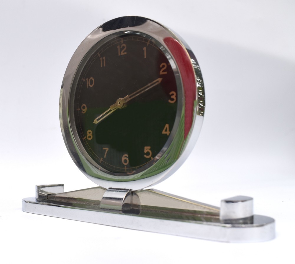 A circular Art Deco desk clock featuring a black dial with gold numerals, mounted on a geometric polished chrome base. View II