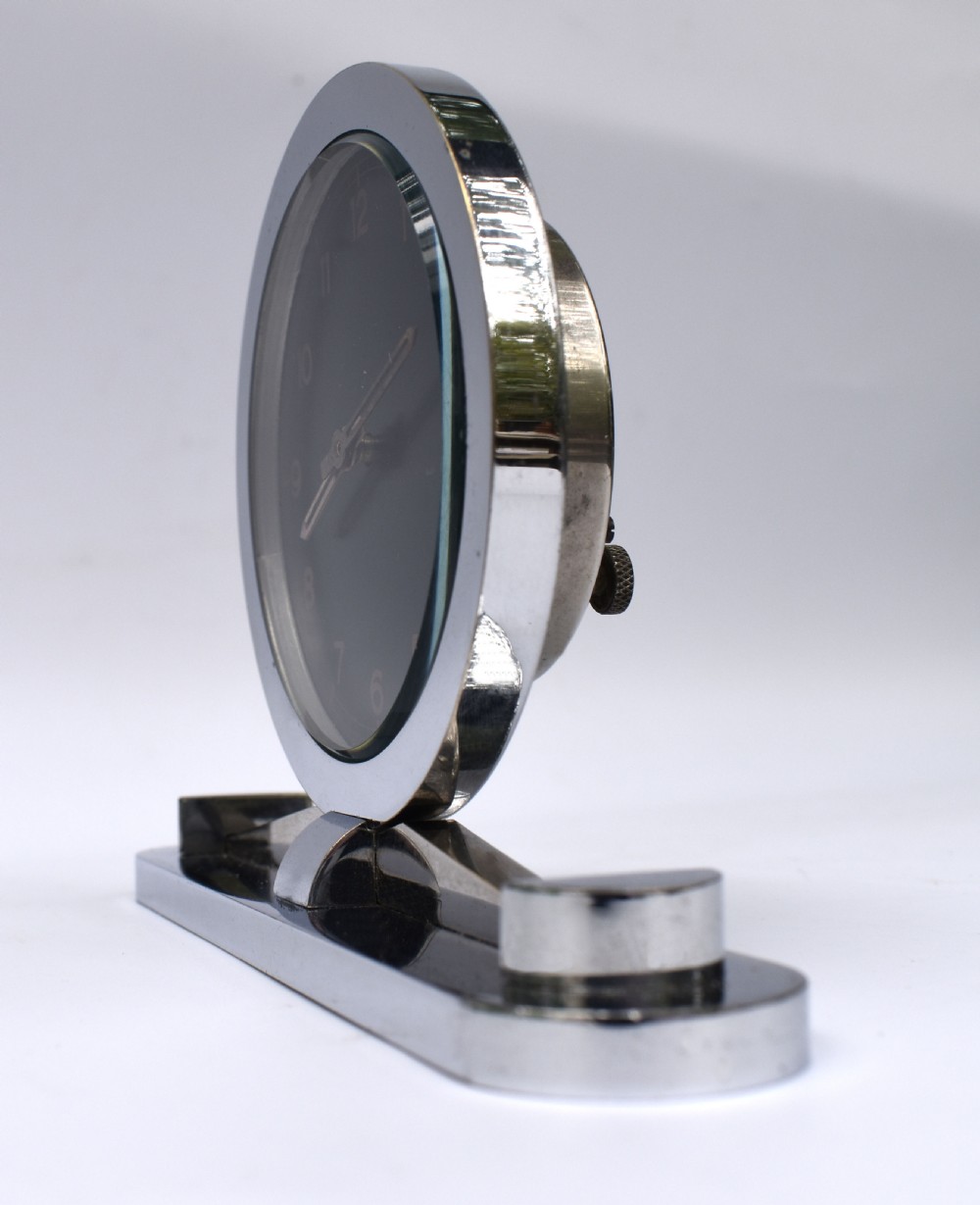 A circular Art Deco desk clock featuring a black dial with gold numerals, mounted on a geometric polished chrome base. View III