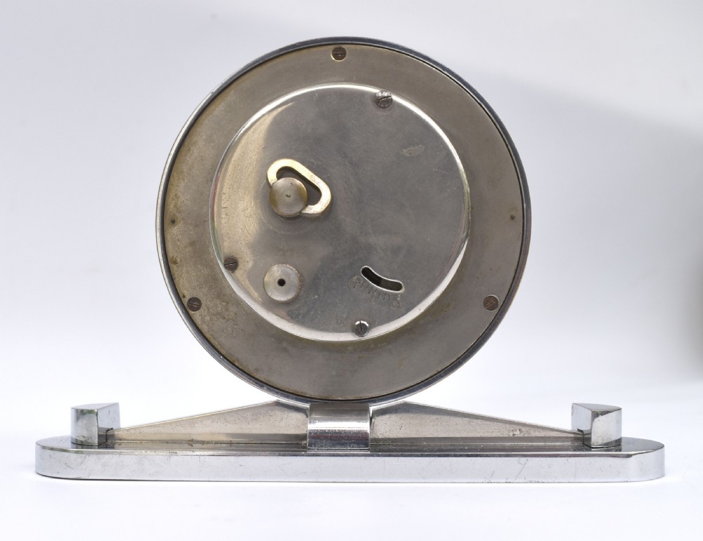 A circular Art Deco desk clock featuring a black dial with gold numerals, mounted on a geometric polished chrome base. View V