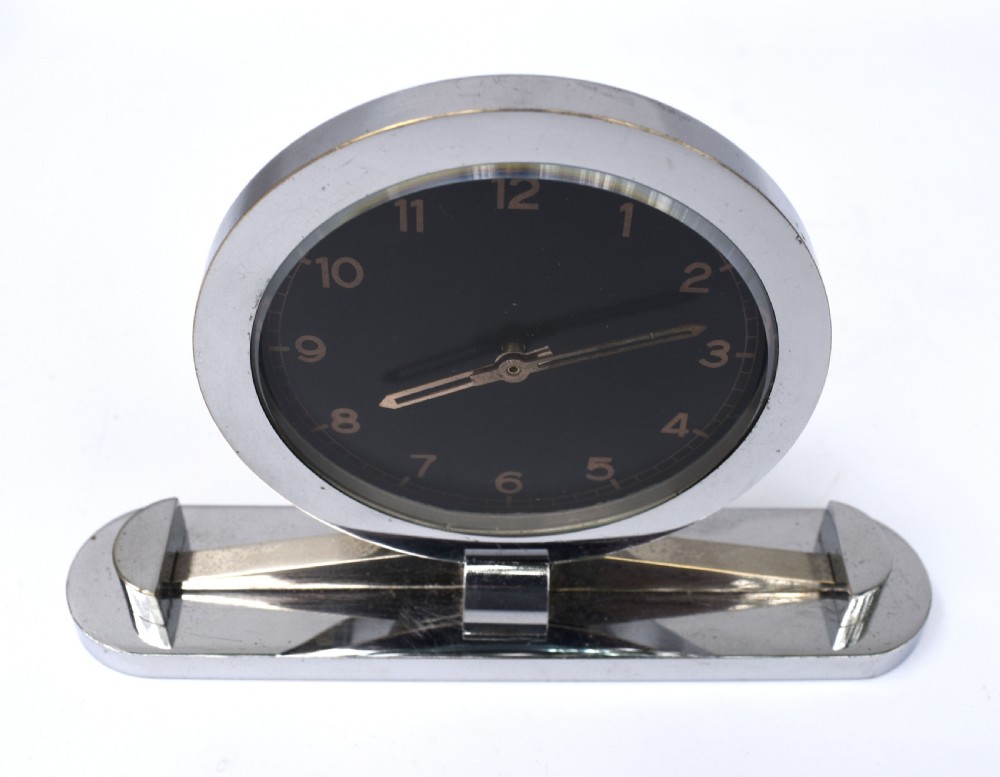 A circular Art Deco desk clock featuring a black dial with gold numerals, mounted on a geometric polished chrome base. View VI