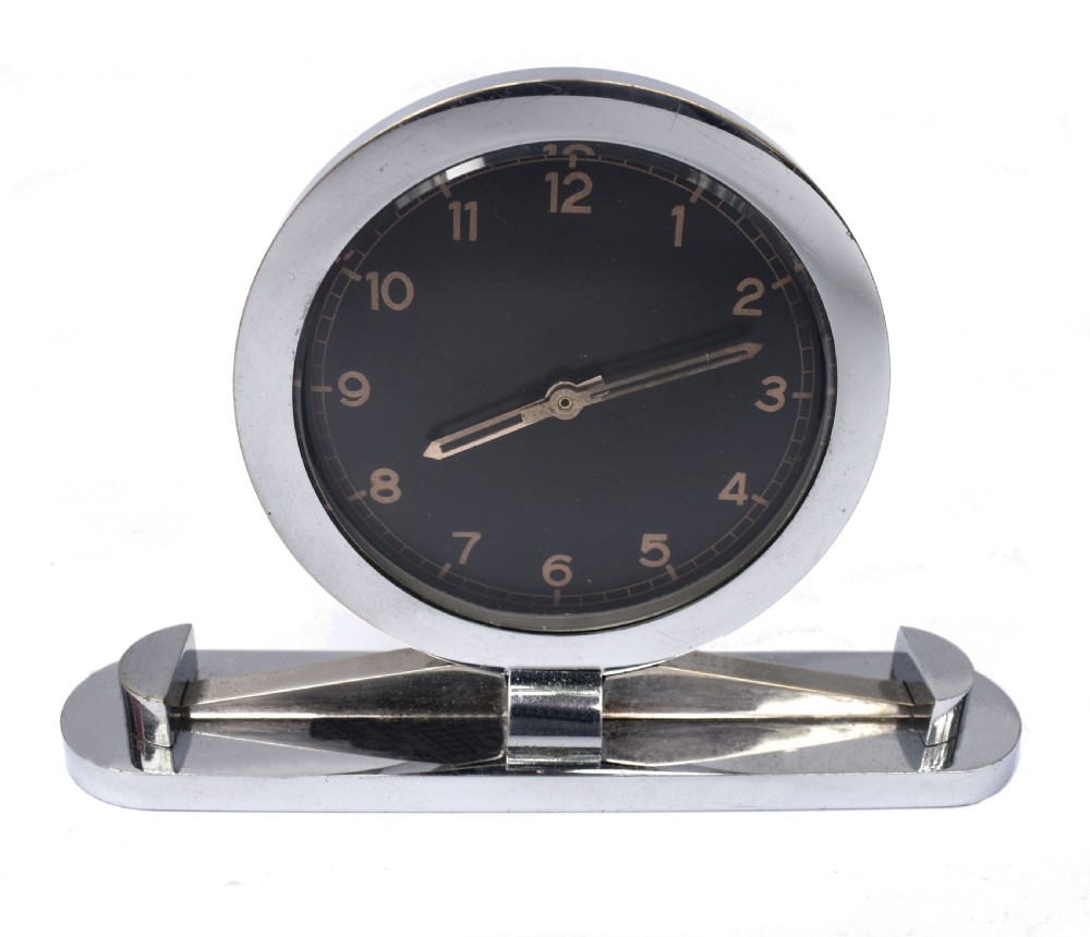 A circular Art Deco desk clock featuring a black dial with gold numerals, mounted on a geometric polished chrome base. View VII