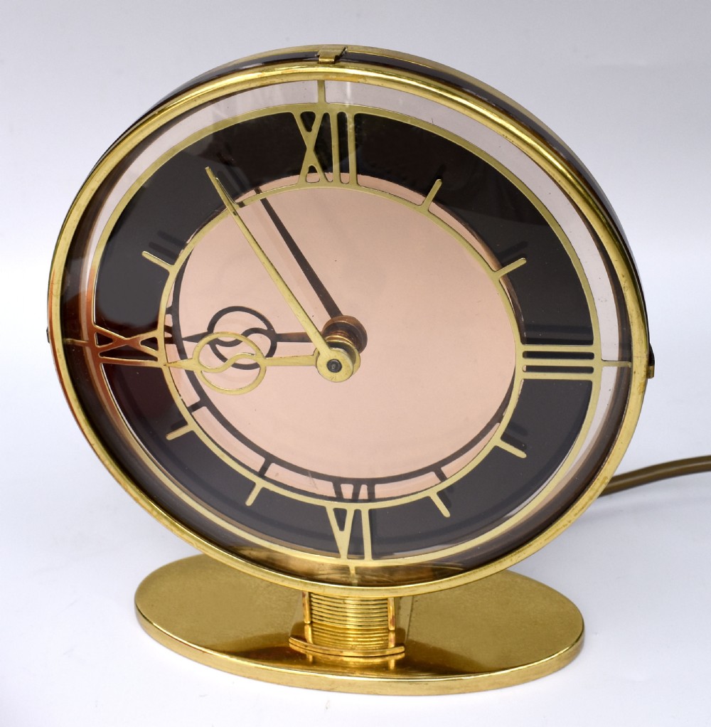 A circular Art Deco brass mantel clock with a pink mirrored dial, Roman numerals, and an oval brass base.