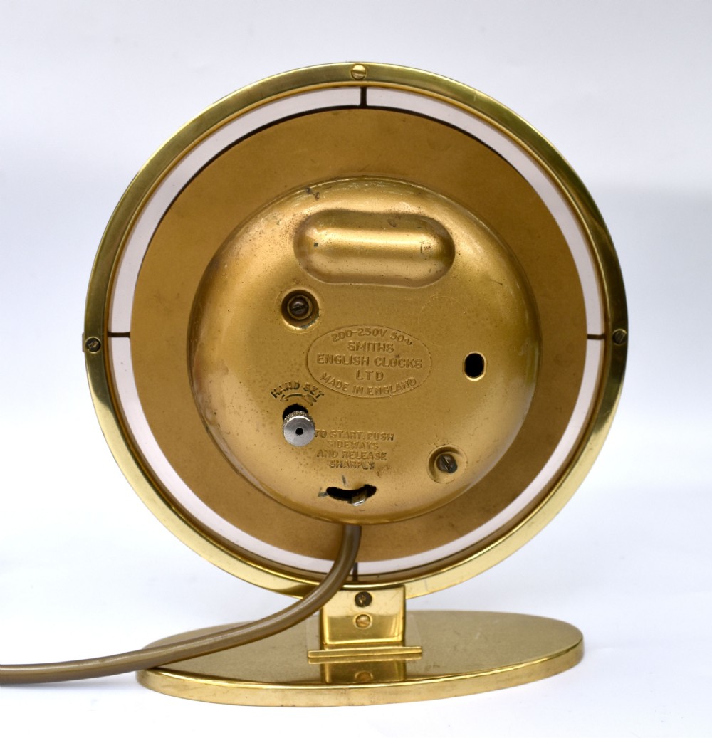 A circular Art Deco brass mantel clock with a pink mirrored dial, Roman numerals, and an oval brass base. View V