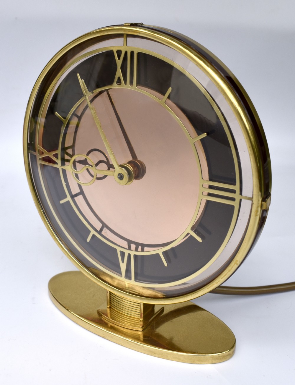 A circular Art Deco brass mantel clock with a pink mirrored dial, Roman numerals, and an oval brass base. View VI