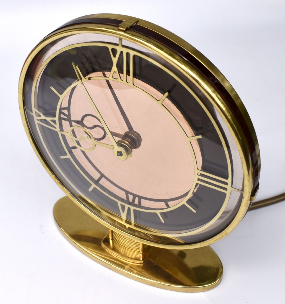 A circular Art Deco brass mantel clock with a pink mirrored dial, Roman numerals, and an oval brass base. View VII