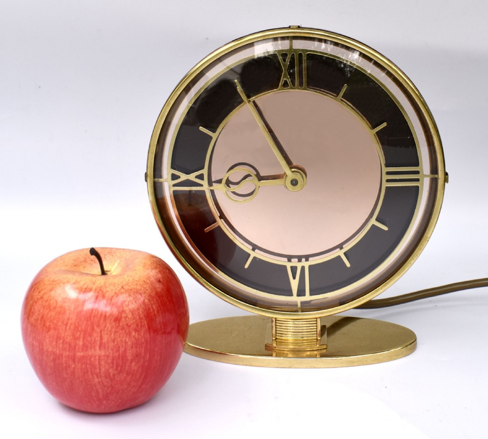 A circular Art Deco brass mantel clock with a pink mirrored dial, Roman numerals, and an oval brass base. View VIII