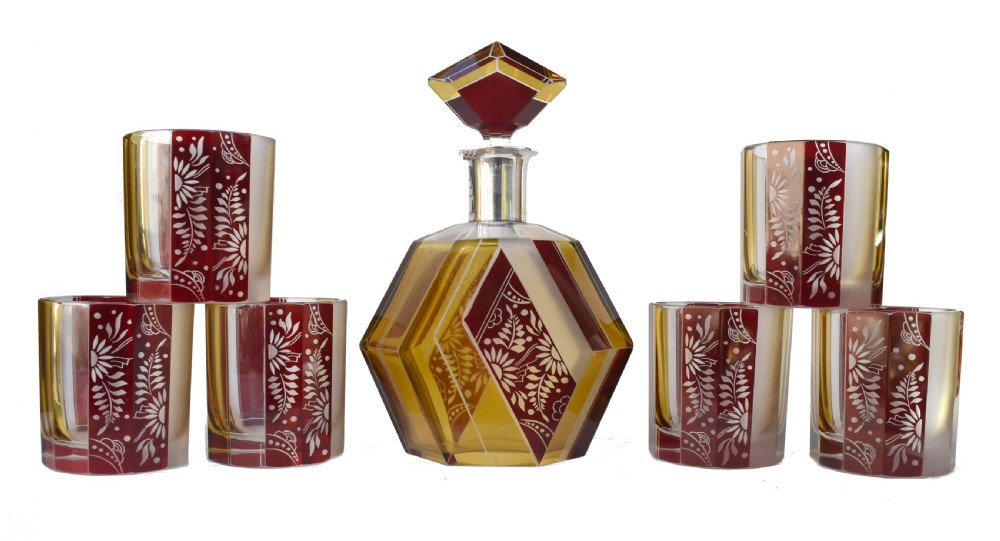 An Art Deco geometric glass decanter with a diamond-shaped stopper surrounded by six matching glass tumblers featuring amber glass and red floral etched panels.