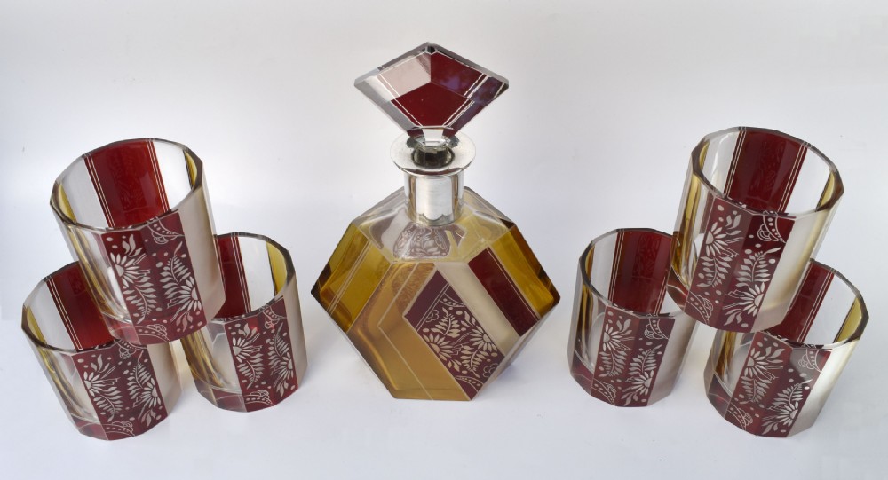 An Art Deco geometric glass decanter with a diamond-shaped stopper surrounded by six matching glass tumblers featuring amber glass and red floral etched panels. View III