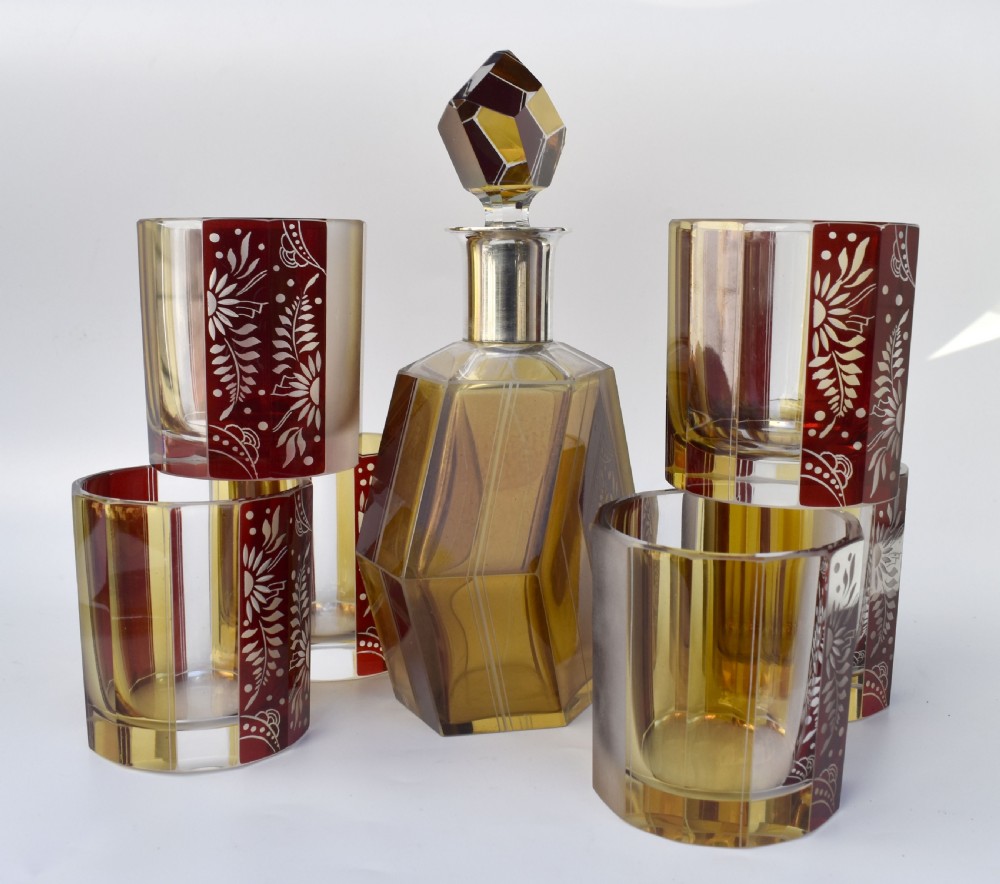 An Art Deco geometric glass decanter with a diamond-shaped stopper surrounded by six matching glass tumblers featuring amber glass and red floral etched panels. View IV