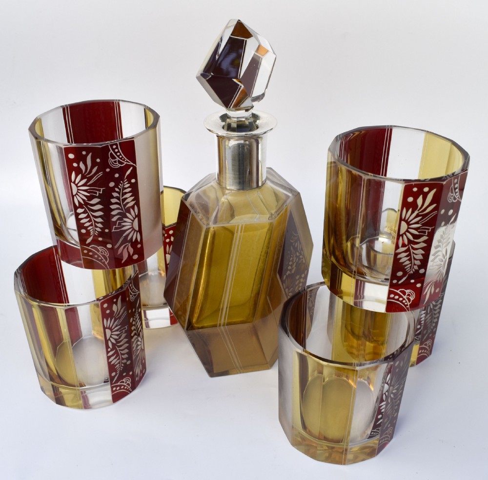 An Art Deco geometric glass decanter with a diamond-shaped stopper surrounded by six matching glass tumblers featuring amber glass and red floral etched panels. View V