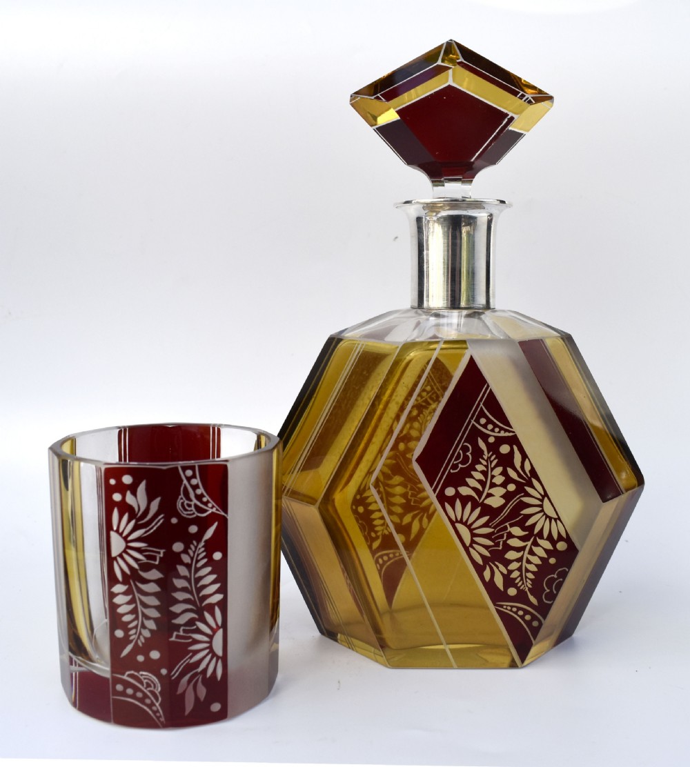 An Art Deco geometric glass decanter with a diamond-shaped stopper surrounded by six matching glass tumblers featuring amber glass and red floral etched panels. View VI