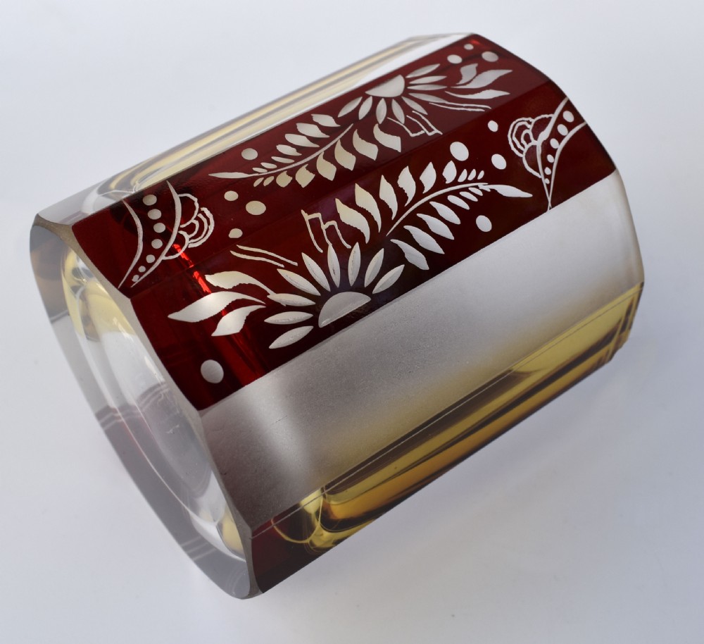 An Art Deco geometric glass decanter with a diamond-shaped stopper surrounded by six matching glass tumblers featuring amber glass and red floral etched panels. View IX