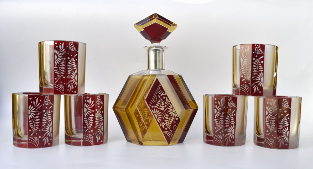 An Art Deco geometric glass decanter with a diamond-shaped stopper surrounded by six matching glass tumblers featuring amber glass and red floral etched panels. View X