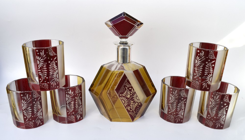 An Art Deco geometric glass decanter with a diamond-shaped stopper surrounded by six matching glass tumblers featuring amber glass and red floral etched panels. View XI