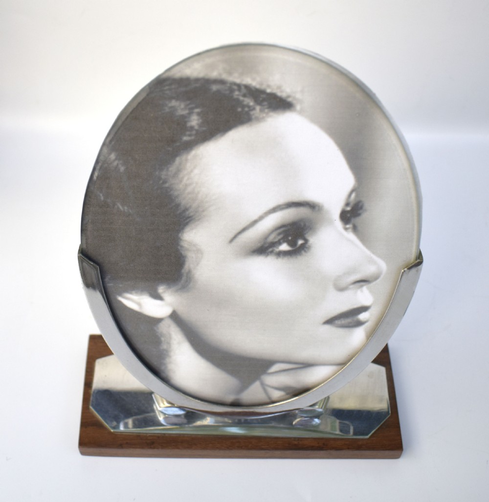 An Art Deco oval chrome photo frame supported by two curved metal brackets on a chrome and dark wood plinth base. View II