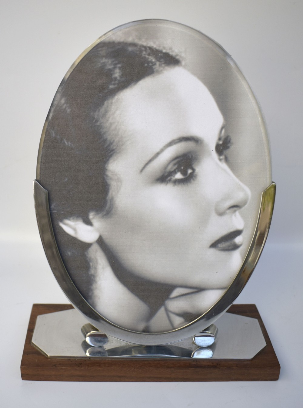 An Art Deco oval chrome photo frame supported by two curved metal brackets on a chrome and dark wood plinth base. View V