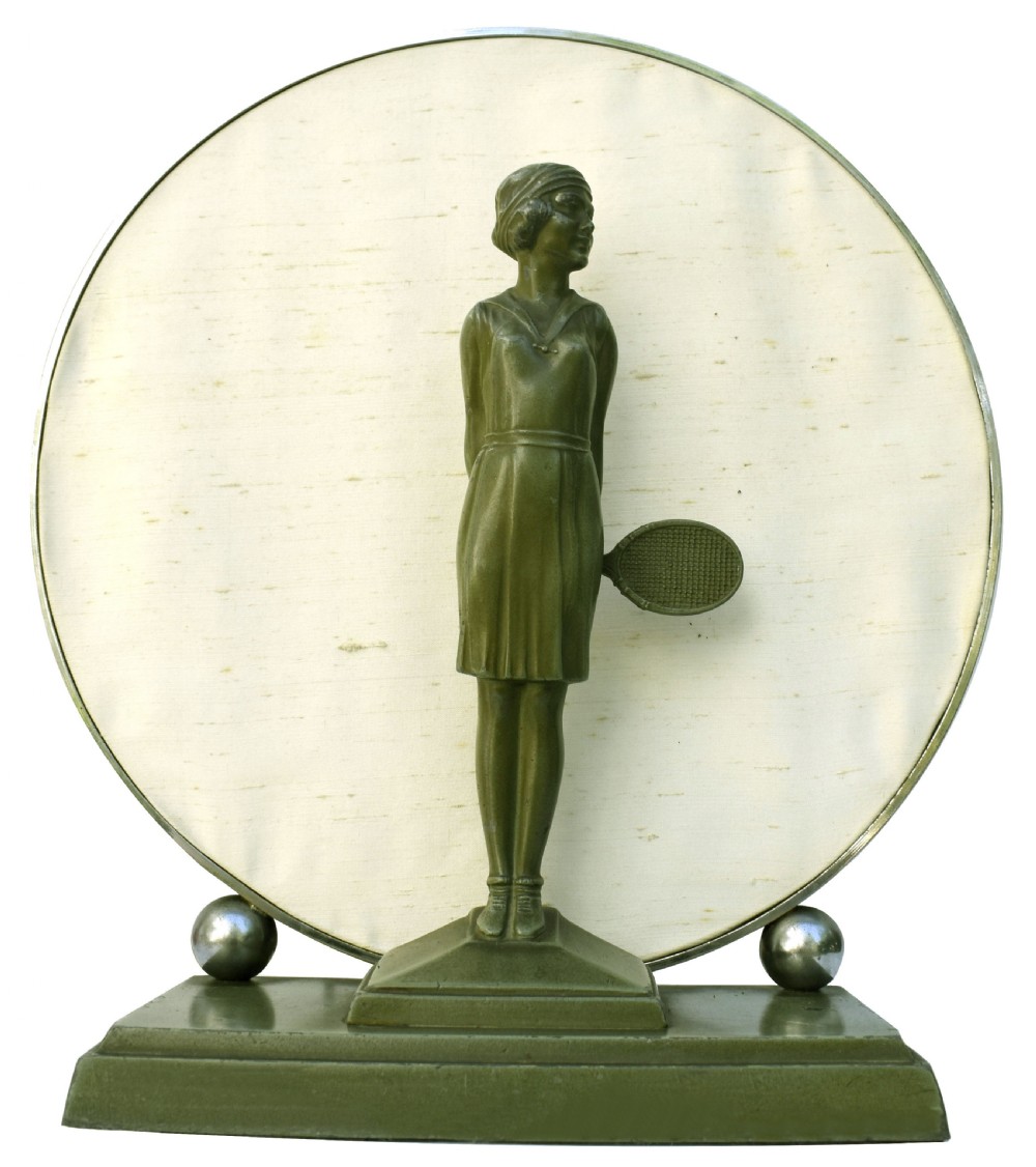 An Art Deco style table lamp featuring a green patinated metal female tennis player figurine standing in front of a circular cream fabric backdrop on a stepped base.