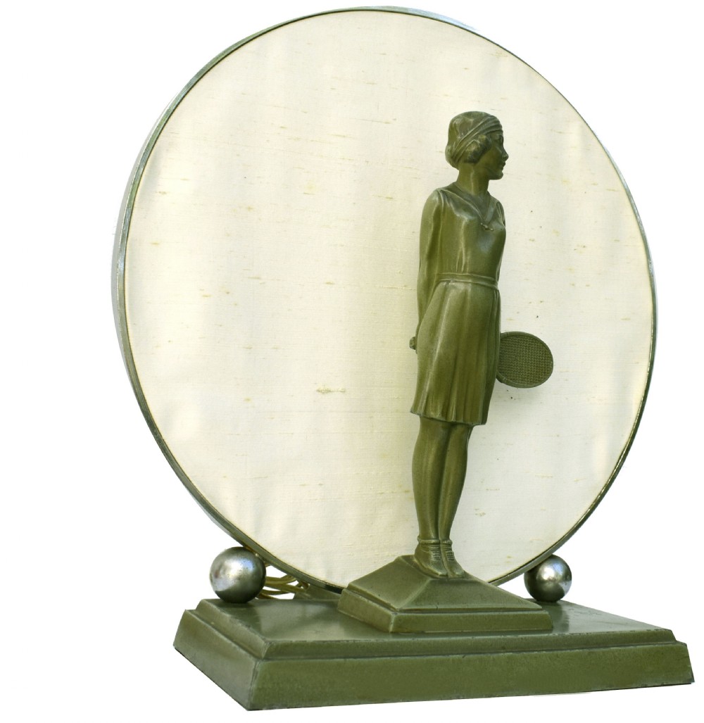 An Art Deco style table lamp featuring a green patinated metal female tennis player figurine standing in front of a circular cream fabric backdrop on a stepped base. View V