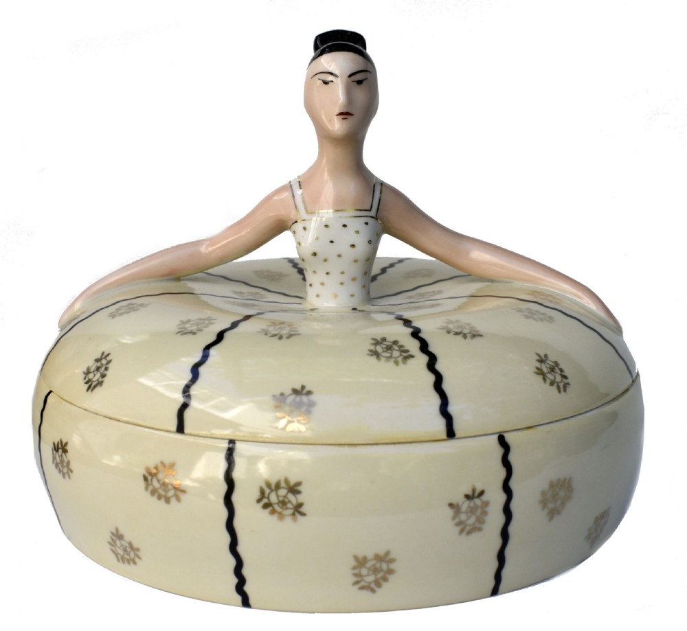 An Art Deco cream ceramic powder bowl with a lid sculpted in the form of a woman wearing a gown decorated with dark vertical wavy stripes and gilt floral motifs.