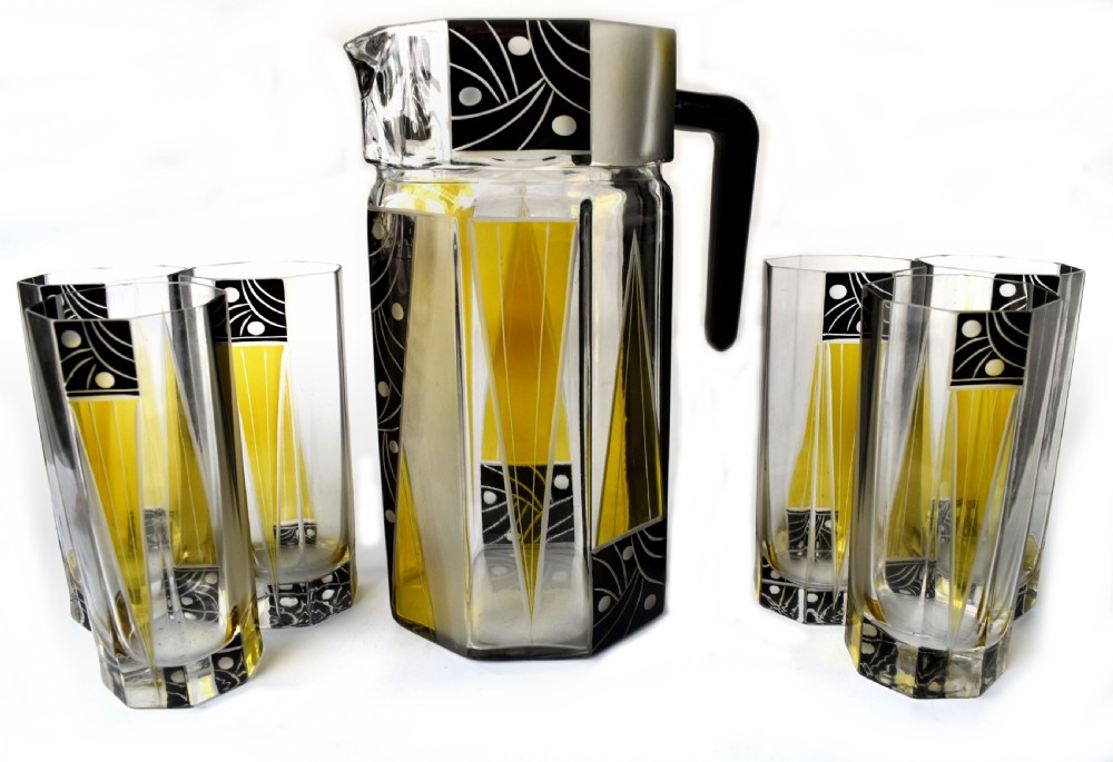 An Art Deco glass drinks set featuring a faceted glass pitcher and six matching tall glasses decorated with yellow and black geometric enamel patterns.