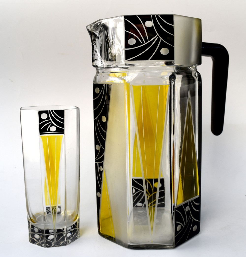 An Art Deco glass drinks set featuring a faceted glass pitcher and six matching tall glasses decorated with yellow and black geometric enamel patterns. View VI