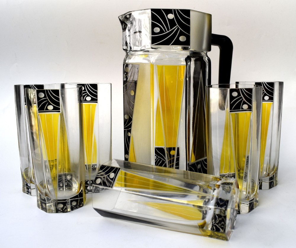 An Art Deco glass drinks set featuring a faceted glass pitcher and six matching tall glasses decorated with yellow and black geometric enamel patterns. View IX