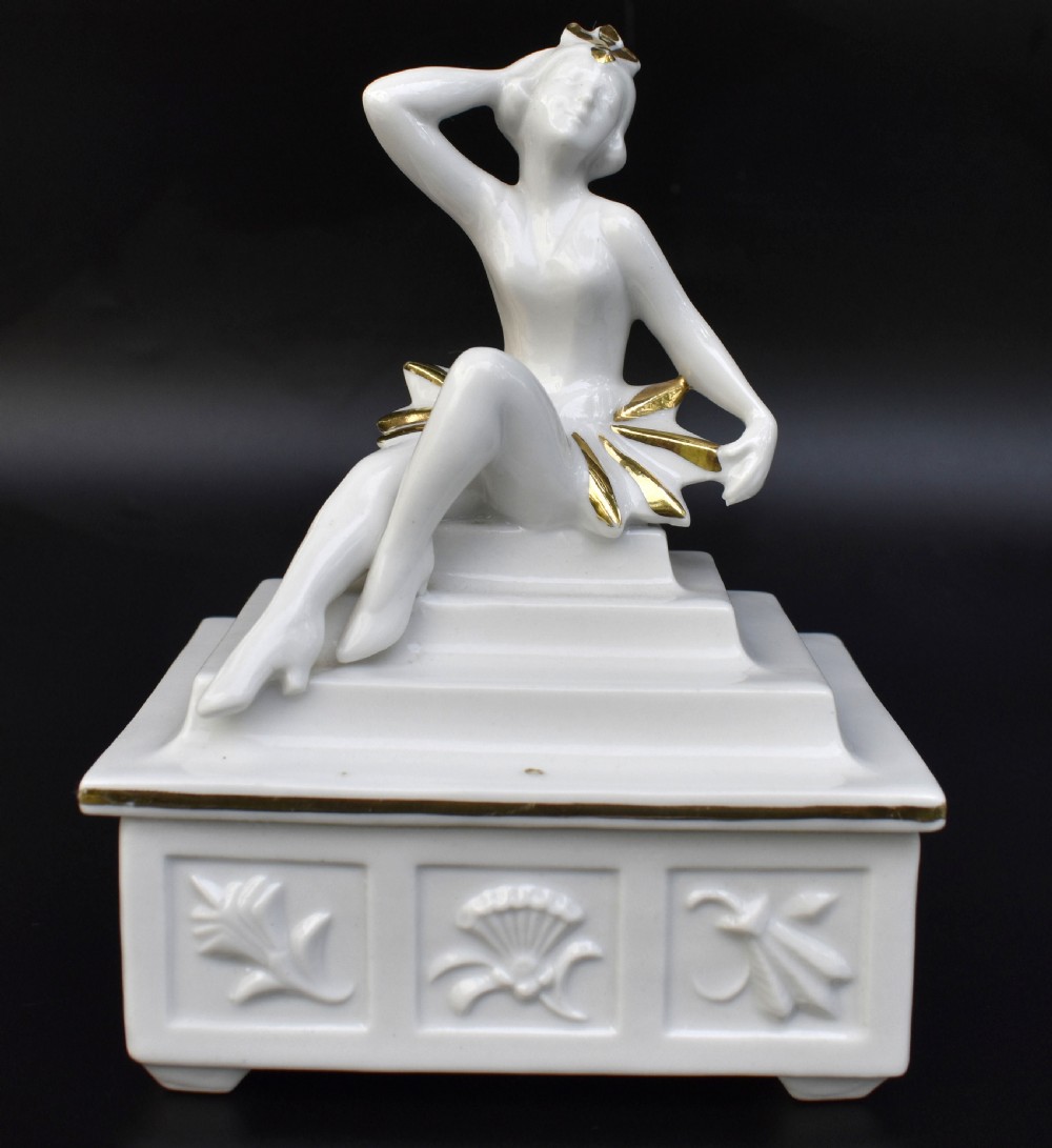A white glazed porcelain Art Deco powder box featuring a lid topped with a seated female figure accented with gold gilding.