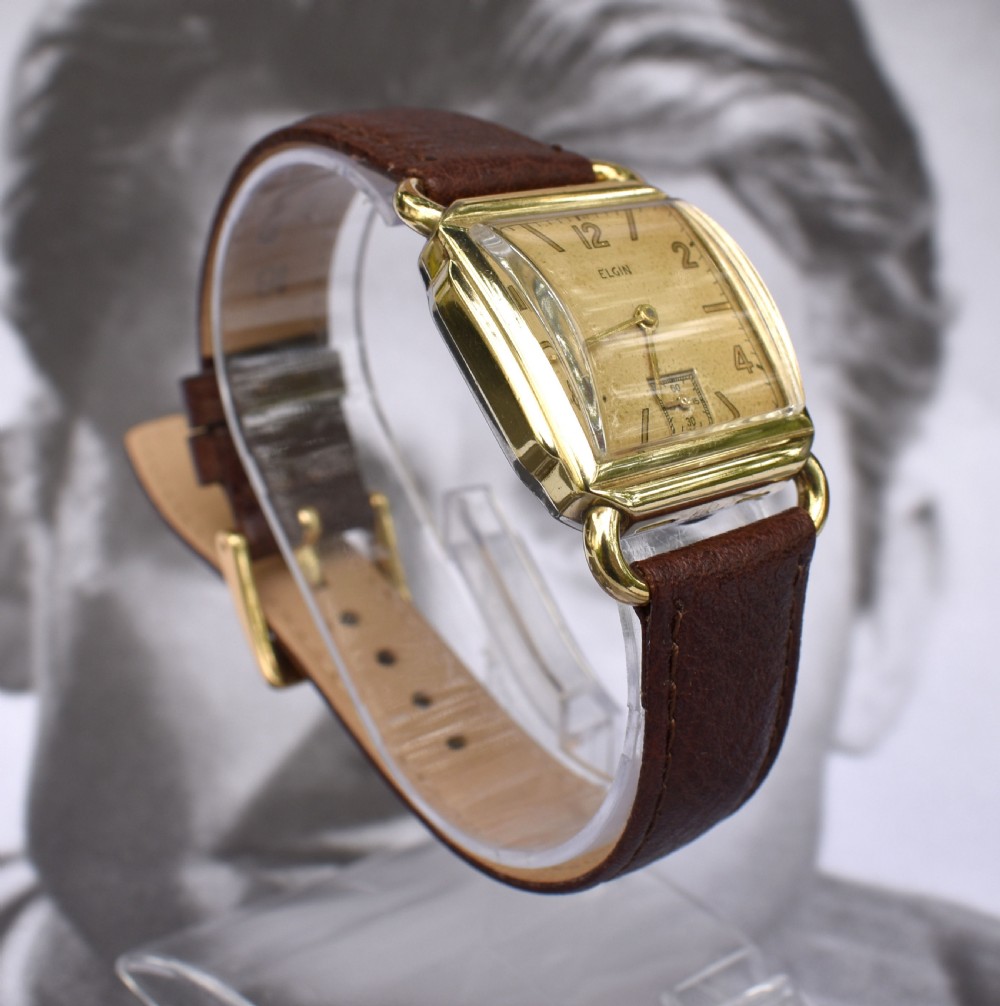 A square gold-filled vintage Elgin gents wristwatch with a champagne dial, gold Arabic numerals, sub-seconds dial, and a dark brown leather strap. View V