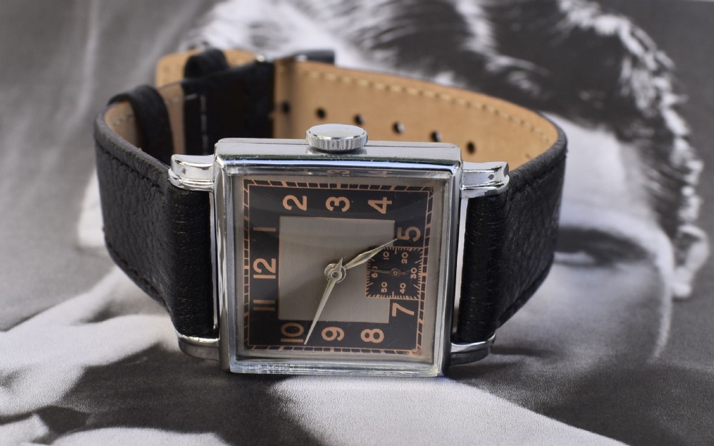 An Art Deco gentlemens wristwatch circa 1935 featuring a rectangular silver-toned case, a black two-tone dial with metallic Arabic numerals, a subdial at six oclock, and a black leather strap. View V