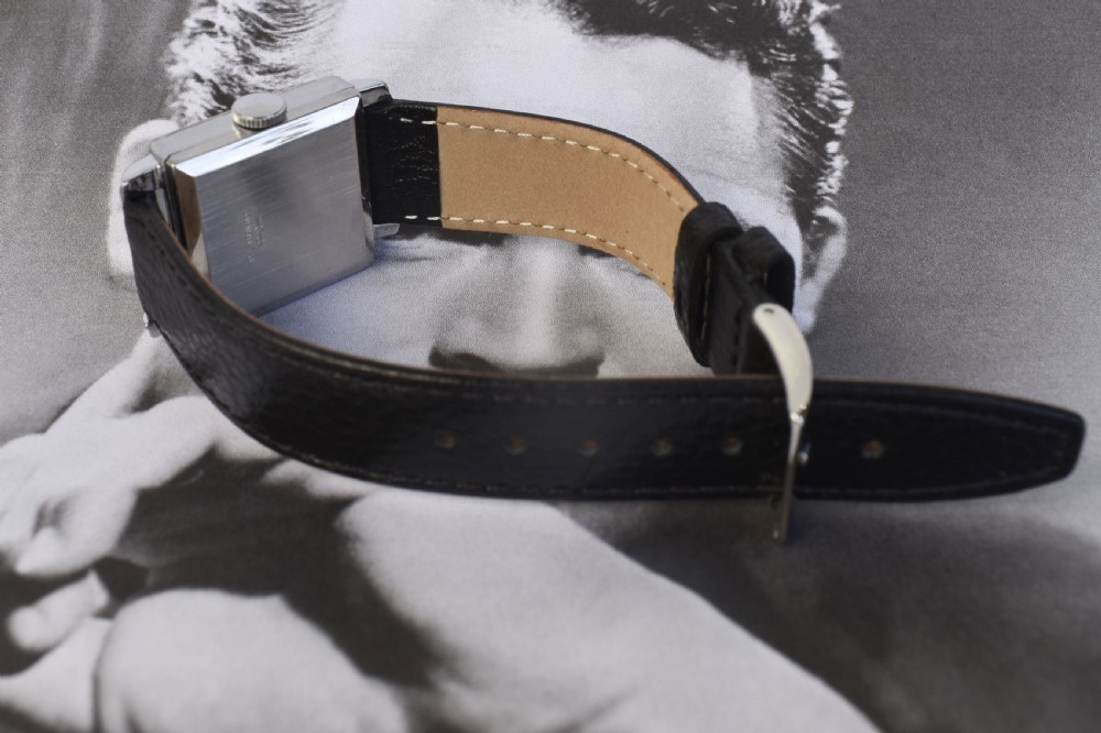An Art Deco gentlemens wristwatch circa 1935 featuring a rectangular silver-toned case, a black two-tone dial with metallic Arabic numerals, a subdial at six oclock, and a black leather strap. View VI