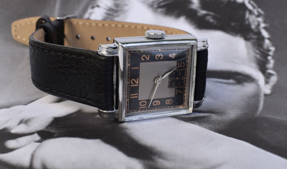 An Art Deco gentlemens wristwatch circa 1935 featuring a rectangular silver-toned case, a black two-tone dial with metallic Arabic numerals, a subdial at six oclock, and a black leather strap. View XII