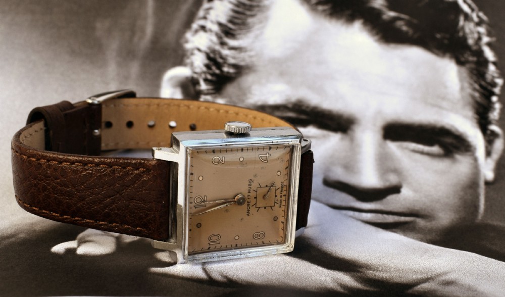An Art Deco gents mechanical wristwatch featuring a square silver-toned case, cream dial with Arabic numerals, and a brown leather strap.