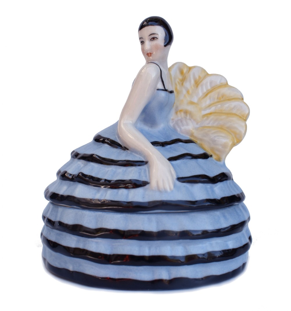 An Art Deco glazed ceramic powder box depicting a female half doll with bobbed black hair and a tiered blue ruffled dress.