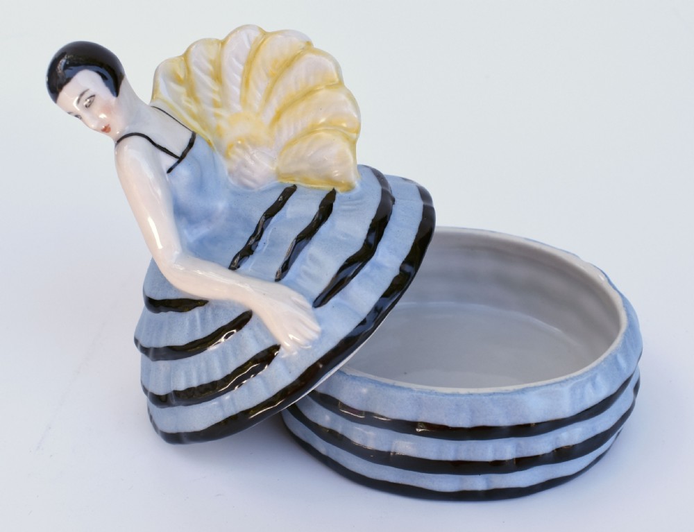 An Art Deco glazed ceramic powder box depicting a female half doll with bobbed black hair and a tiered blue ruffled dress. View VI