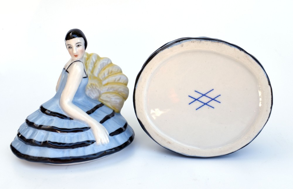 An Art Deco glazed ceramic powder box depicting a female half doll with bobbed black hair and a tiered blue ruffled dress. View VIII