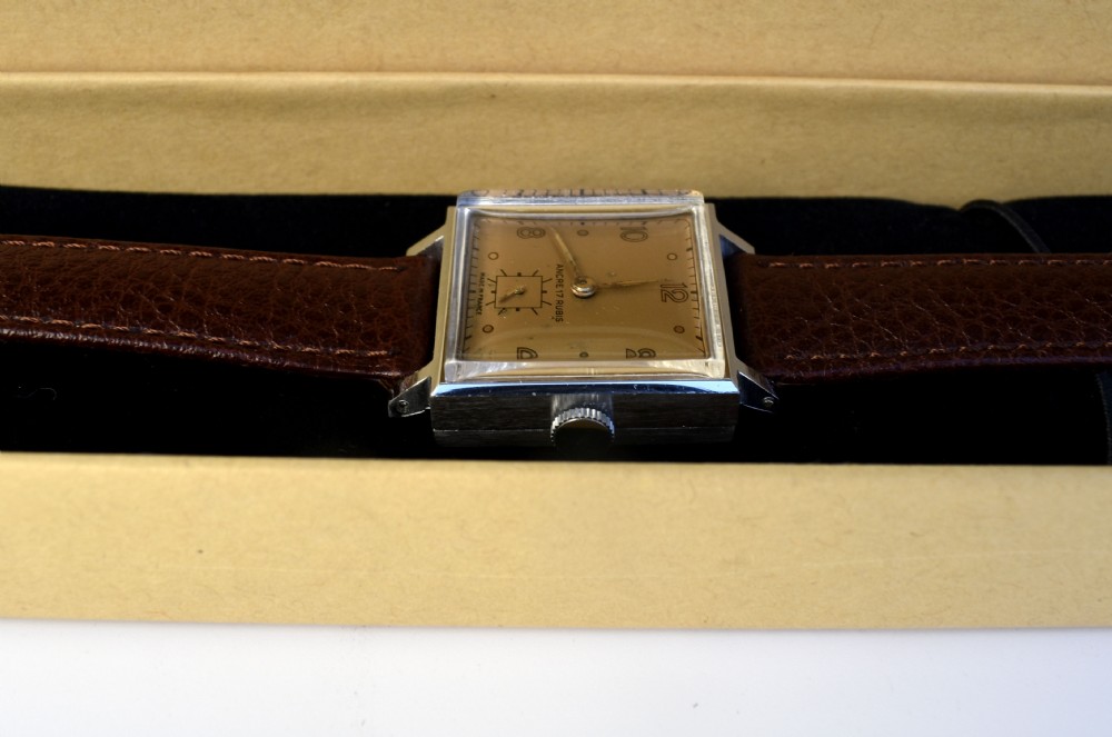 An Art Deco gents mechanical wristwatch featuring a square silver-toned case, cream dial with Arabic numerals, and a brown leather strap. View VII