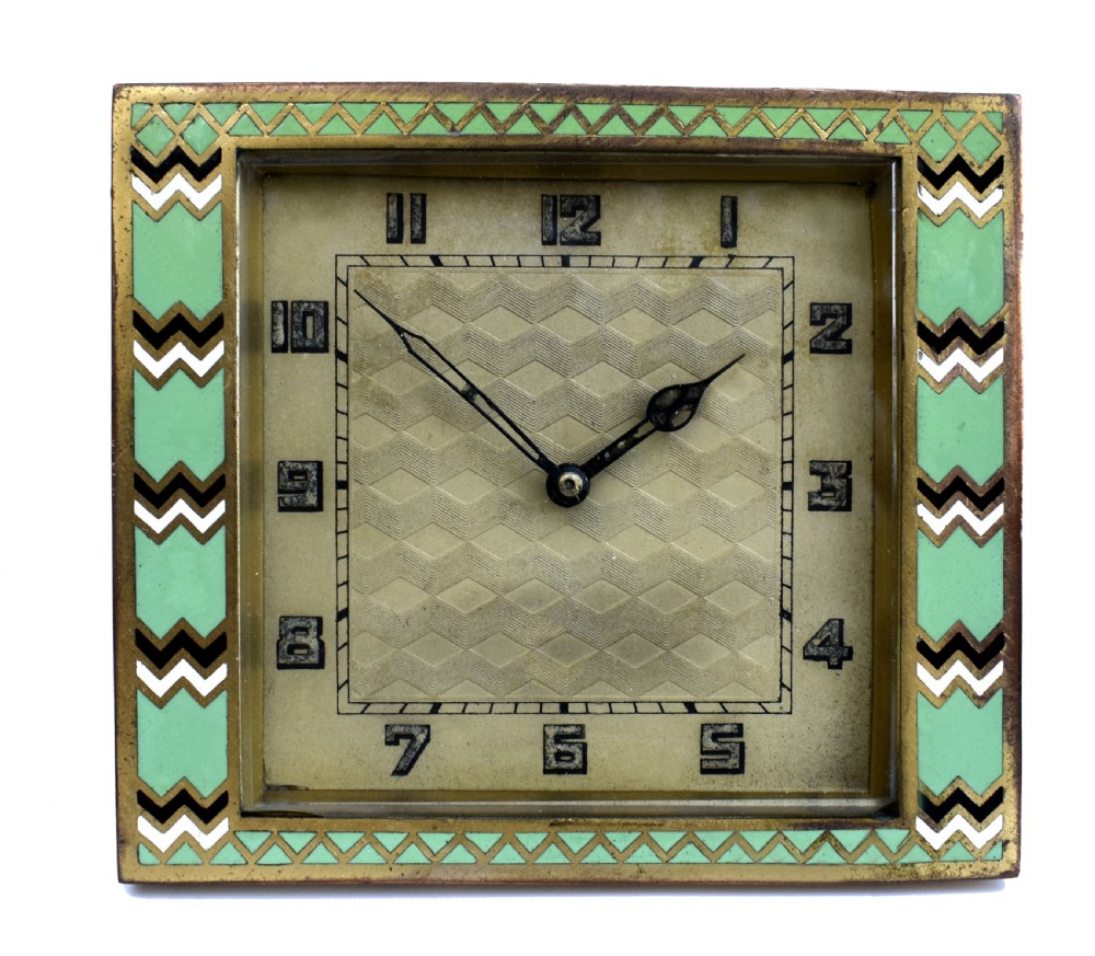 A square Art Deco clock featuring a green, white, and black chevron enamel border surrounding a geometric patterned metal dial with Arabic numerals.
